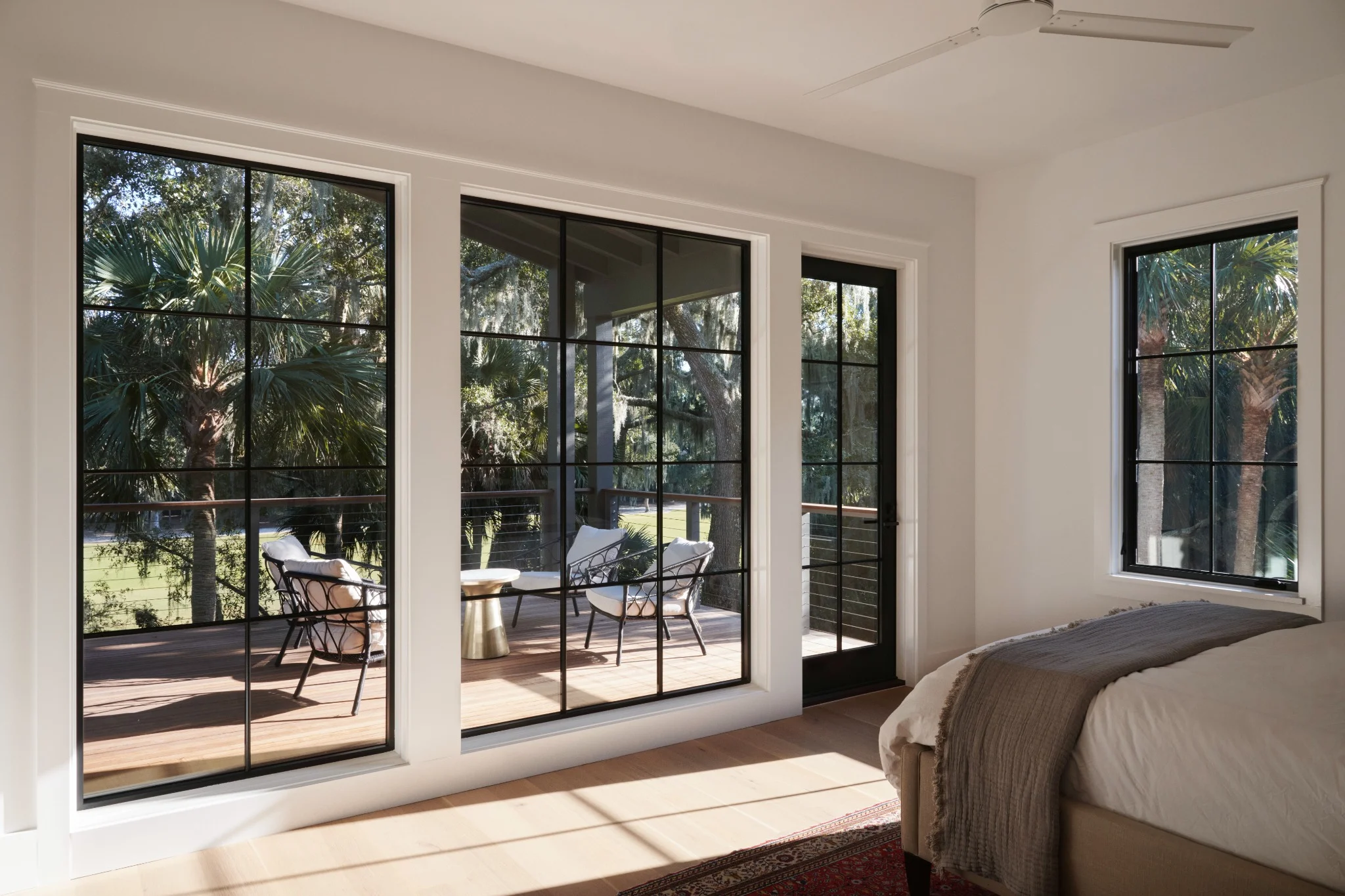 A modern bedroom featuring large black grid-frame windows and a glass door opening to a sunlit deck with lounge chairs