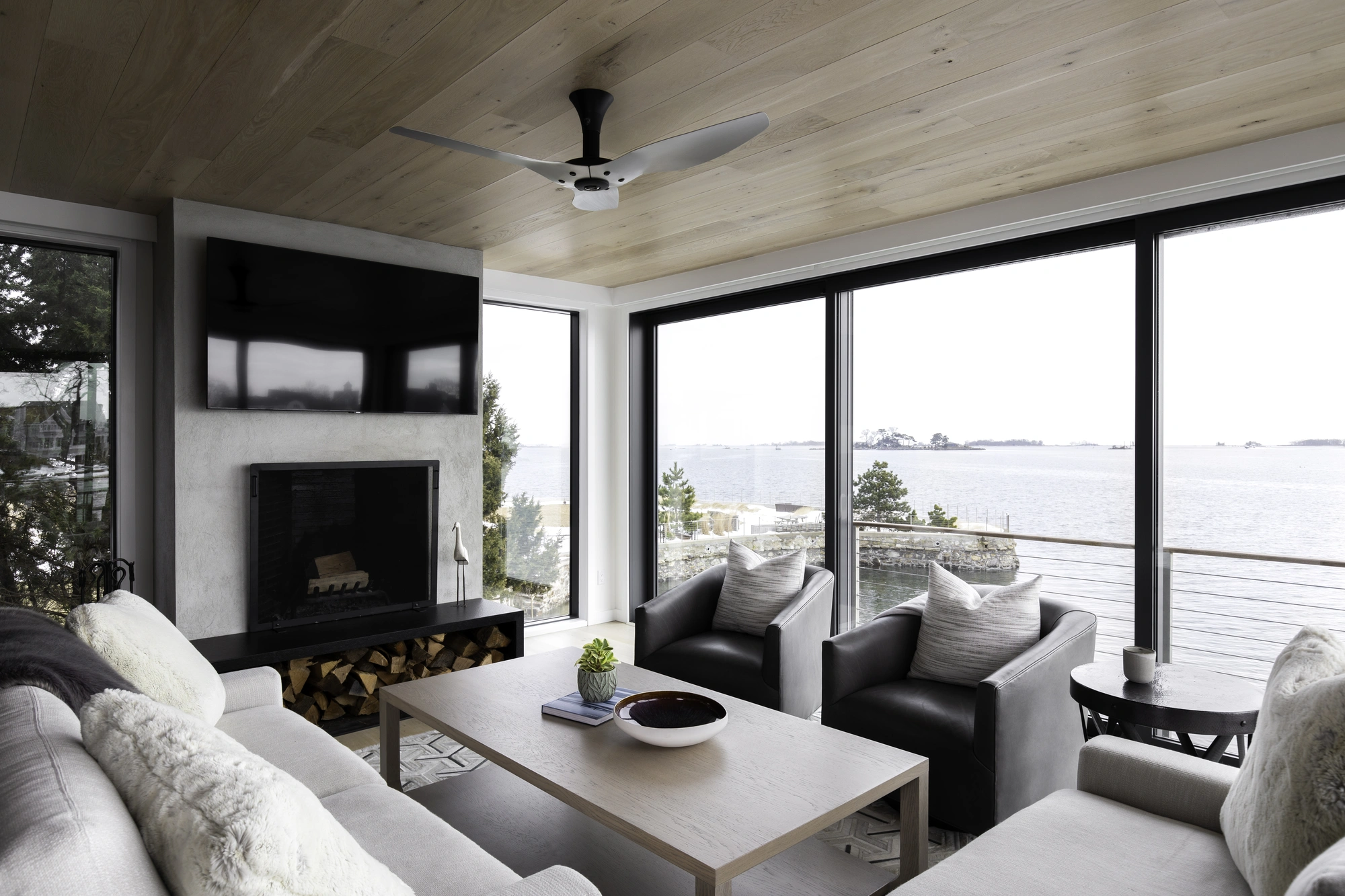 Modern sunroom with floor-to-ceiling windows overlooking the water, featuring a light wood ceiling, fireplace, and cozy seating with scenic views