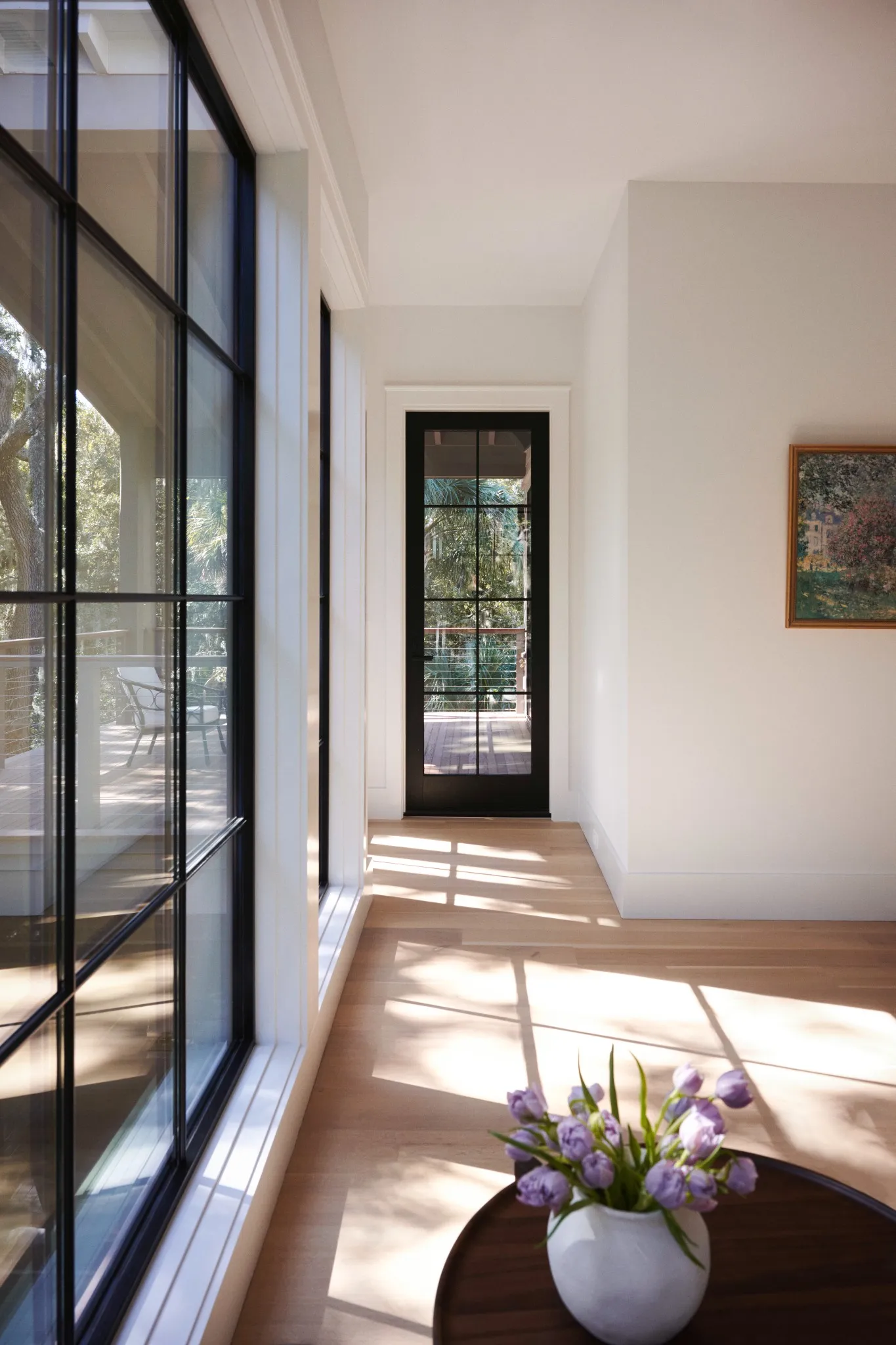 Bright hallway with black-trimmed grid windows and a glass door leading to a deck, showcasing natural light, wood floors, and a vase of purple tulips on a round table