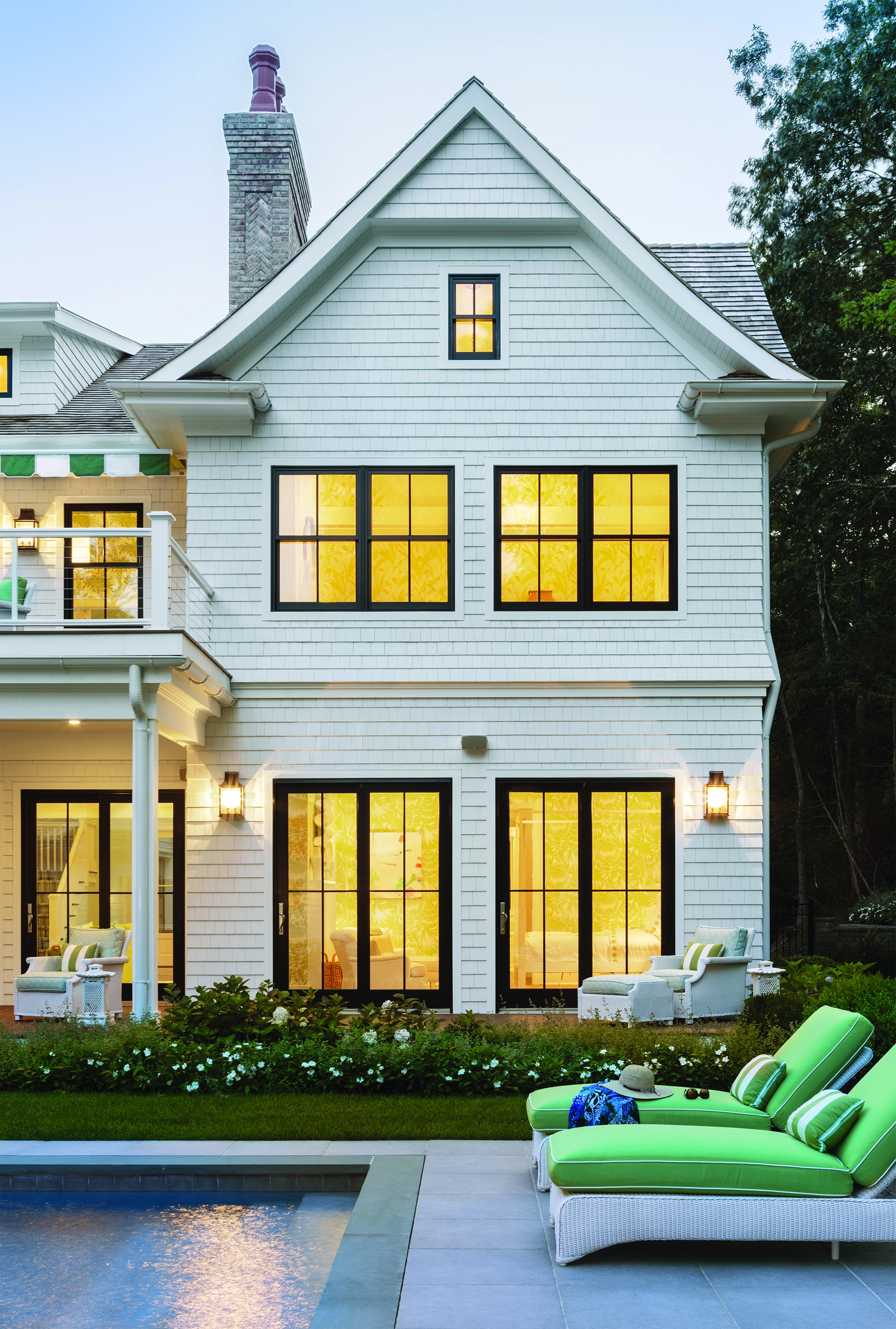 Exterior view of a white two-story home with black-trimmed double-hung windows and French doors, glowing warmly at dusk, with green poolside lounge chairs in the foreground