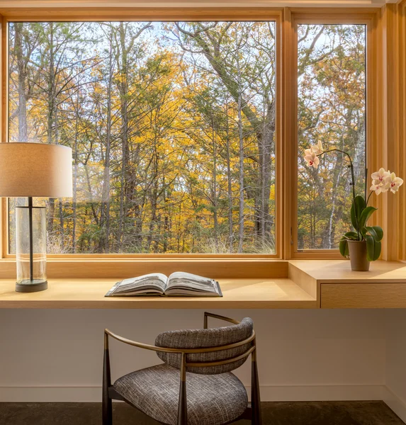 Image of interior wood-framed window overlooking the forest with desk and lamp