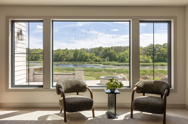 Image of black-framed windows overlooking a pond with two chairs and a side table
